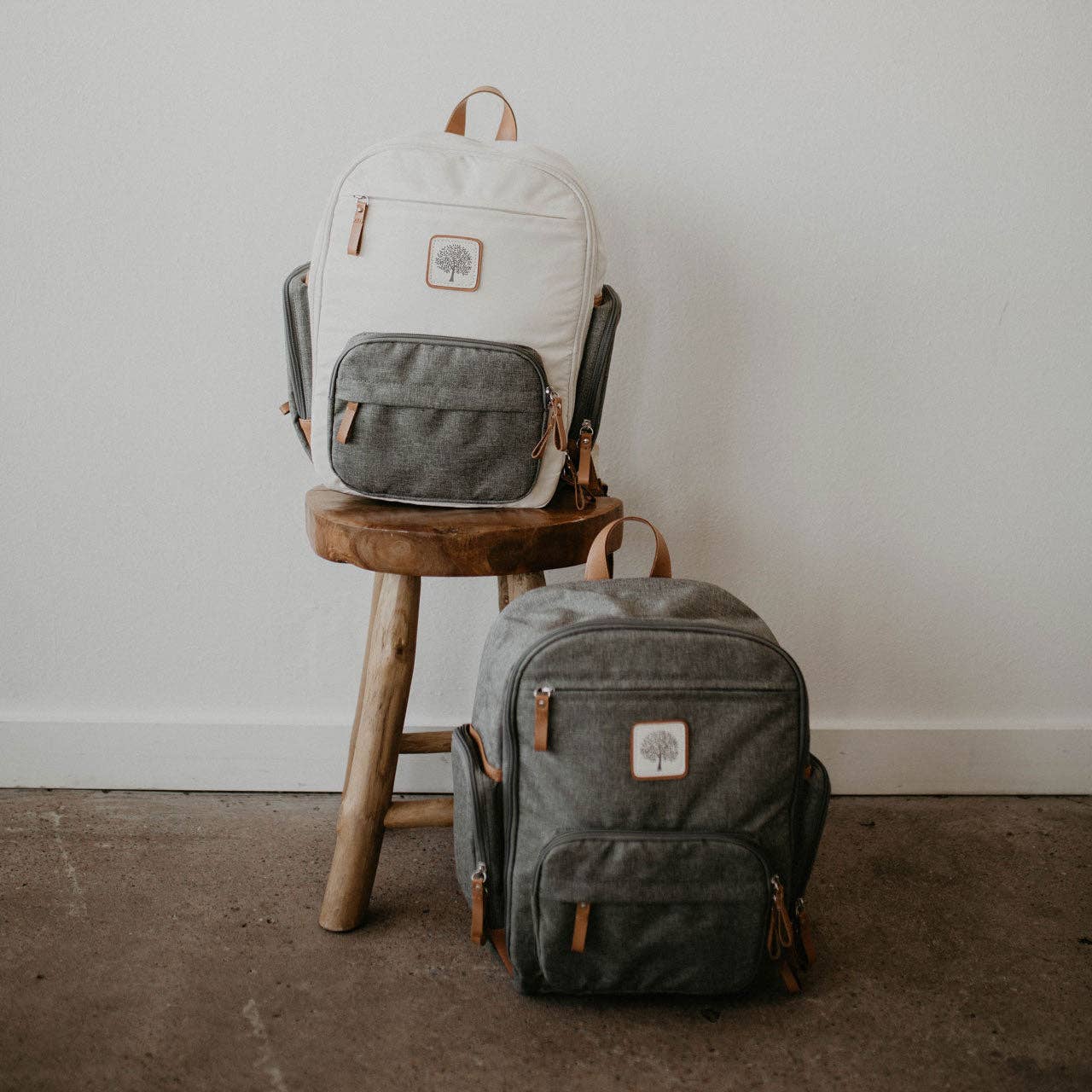 Two backpacks, one white and one gray, on a wooden stool and floor.