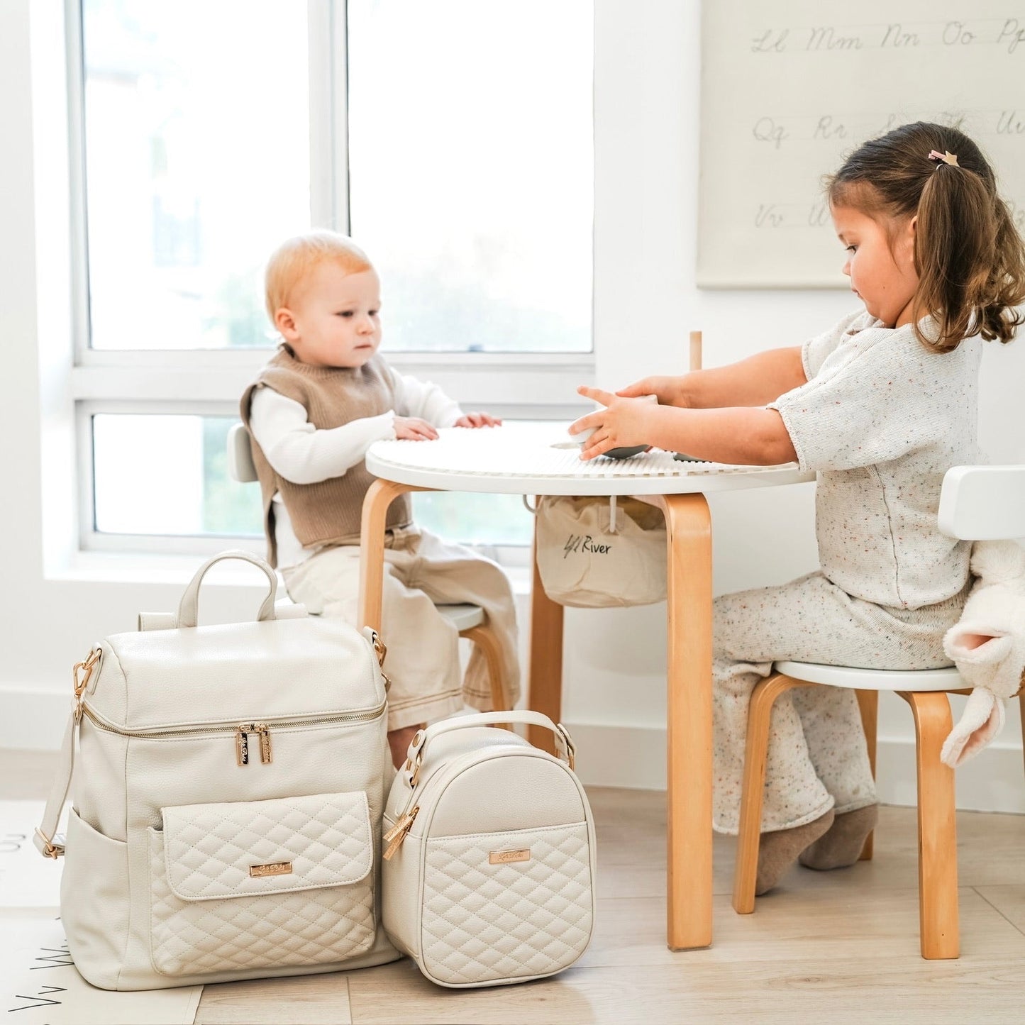 Two children sitting at a table with beige bags on the floor.
