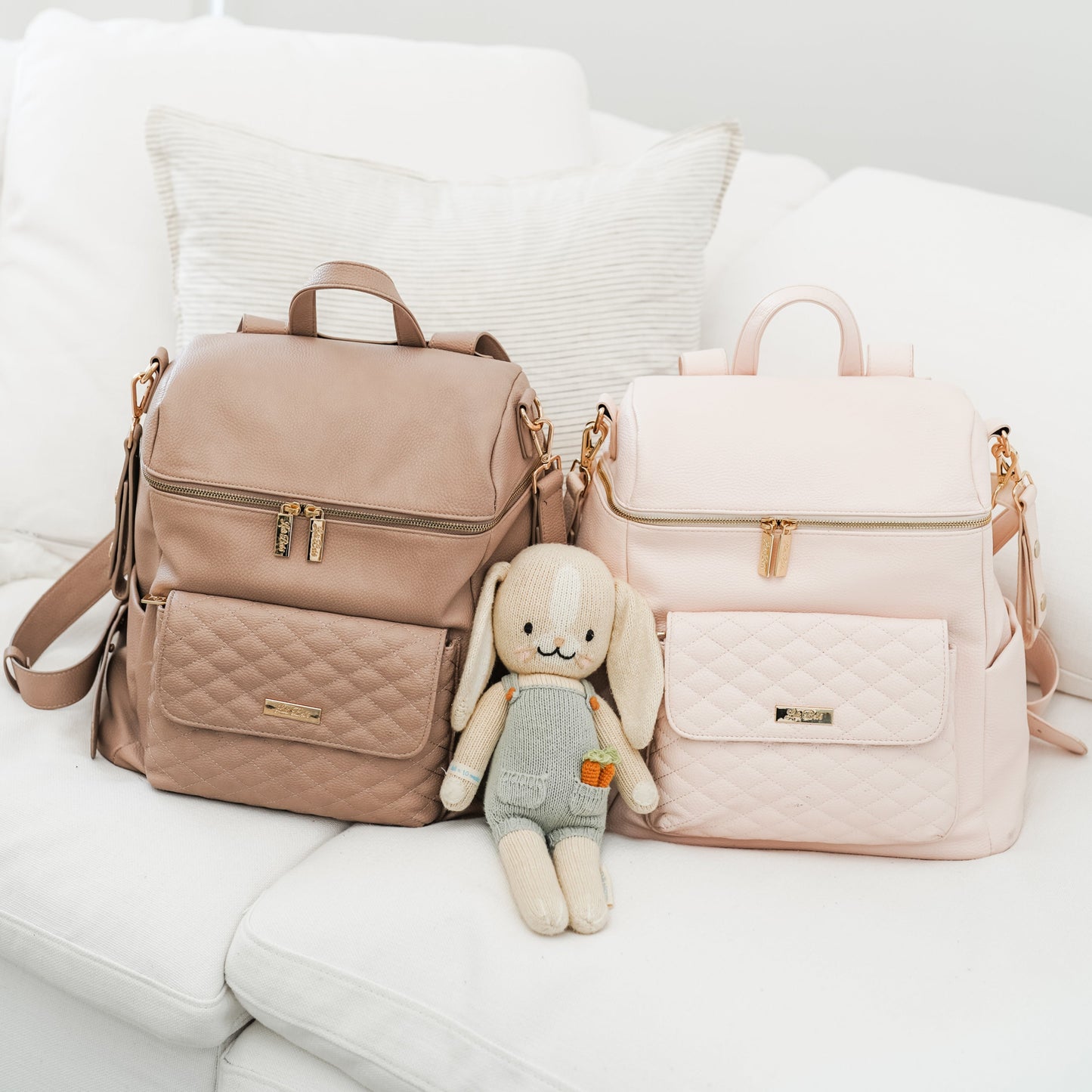 Two backpacks, one beige and one pink, on a white surface with a plush toy between them.