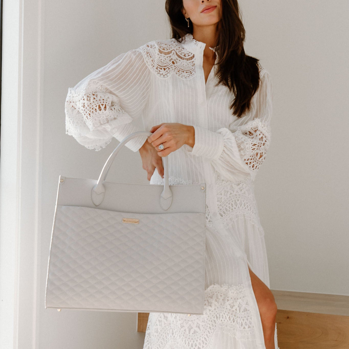 Woman in a white lace dress holding a light gray handbag against a neutral background