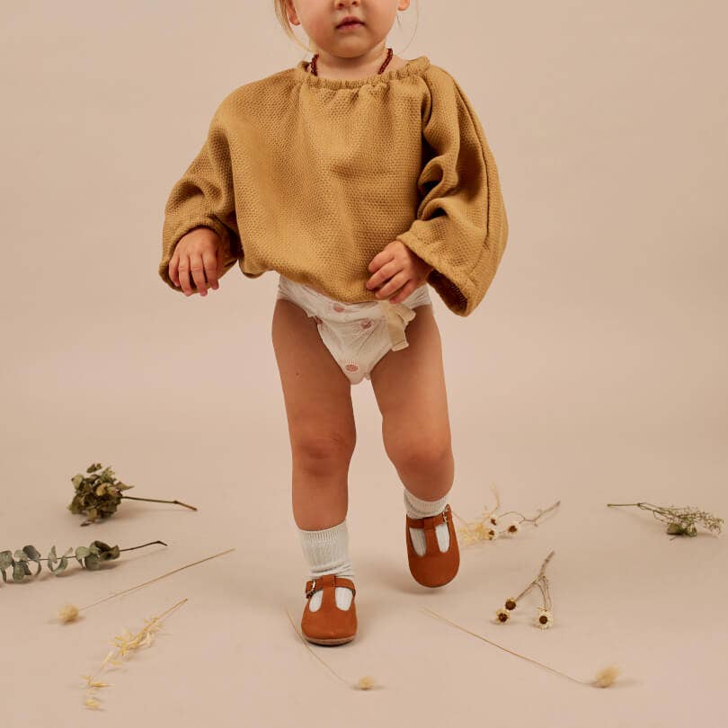 Child wearing a brown blouse and brown shoes with dried flowers on a neutral background.