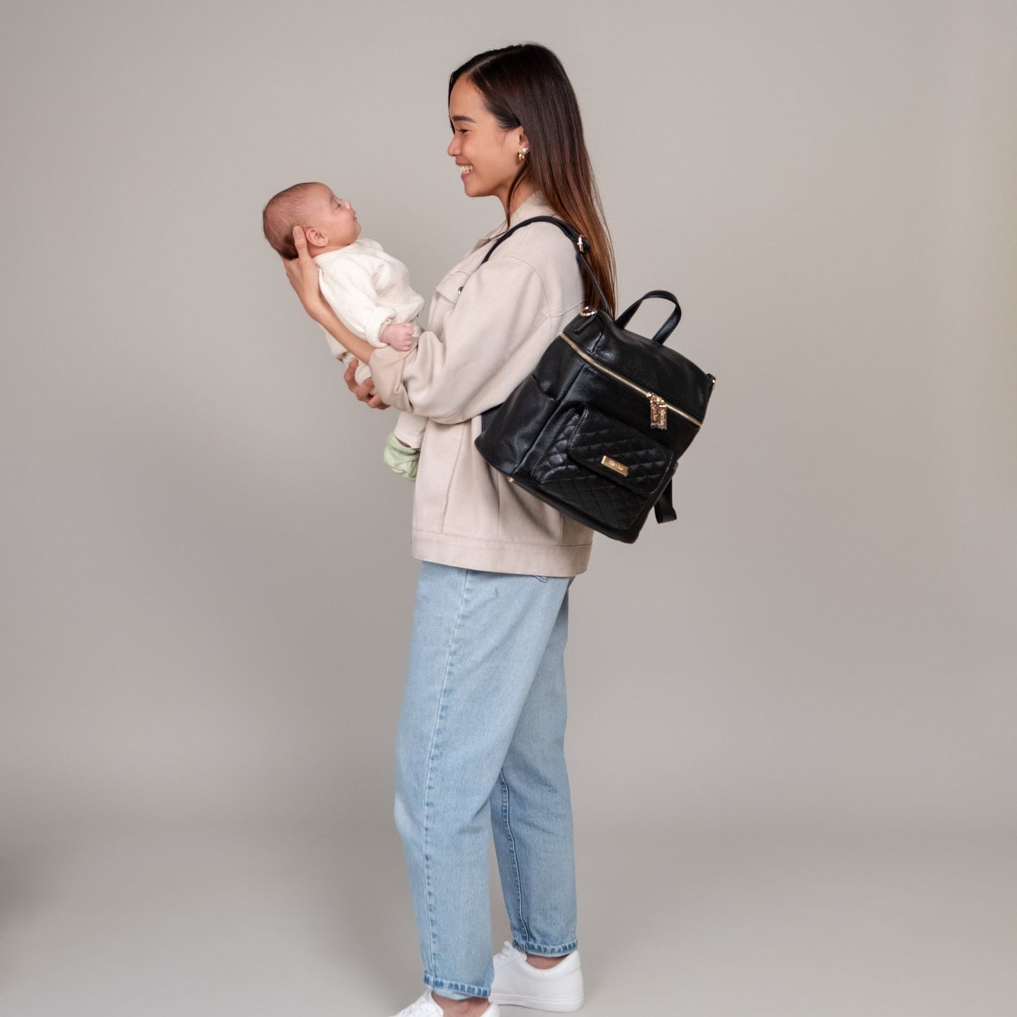 Woman holding a baby and carrying a black backpack against a plain background