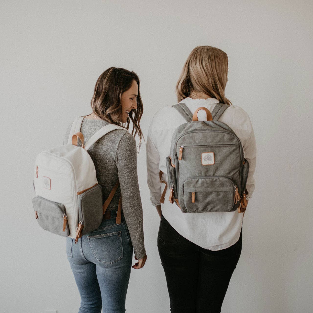Two women wearing backpacks, one white and one gray, against a plain background.
