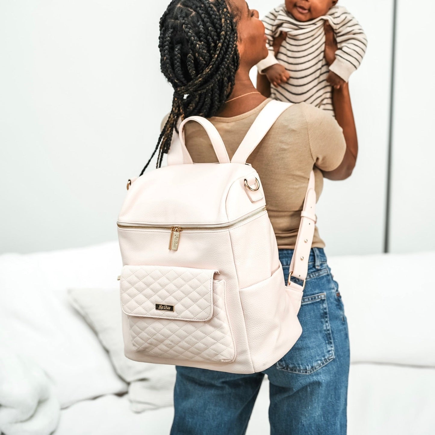 Woman holding a baby and wearing a light pink quilted backpack indoors.