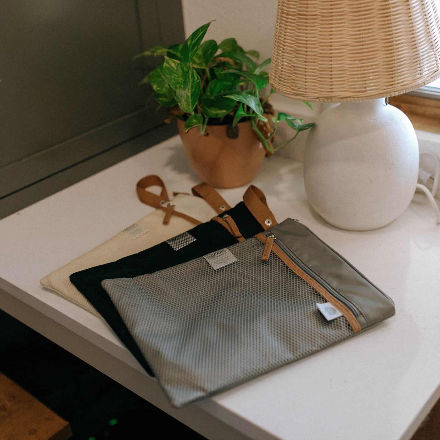 Gray pouch with brown straps on a white table, with a lamp and plant in the background.