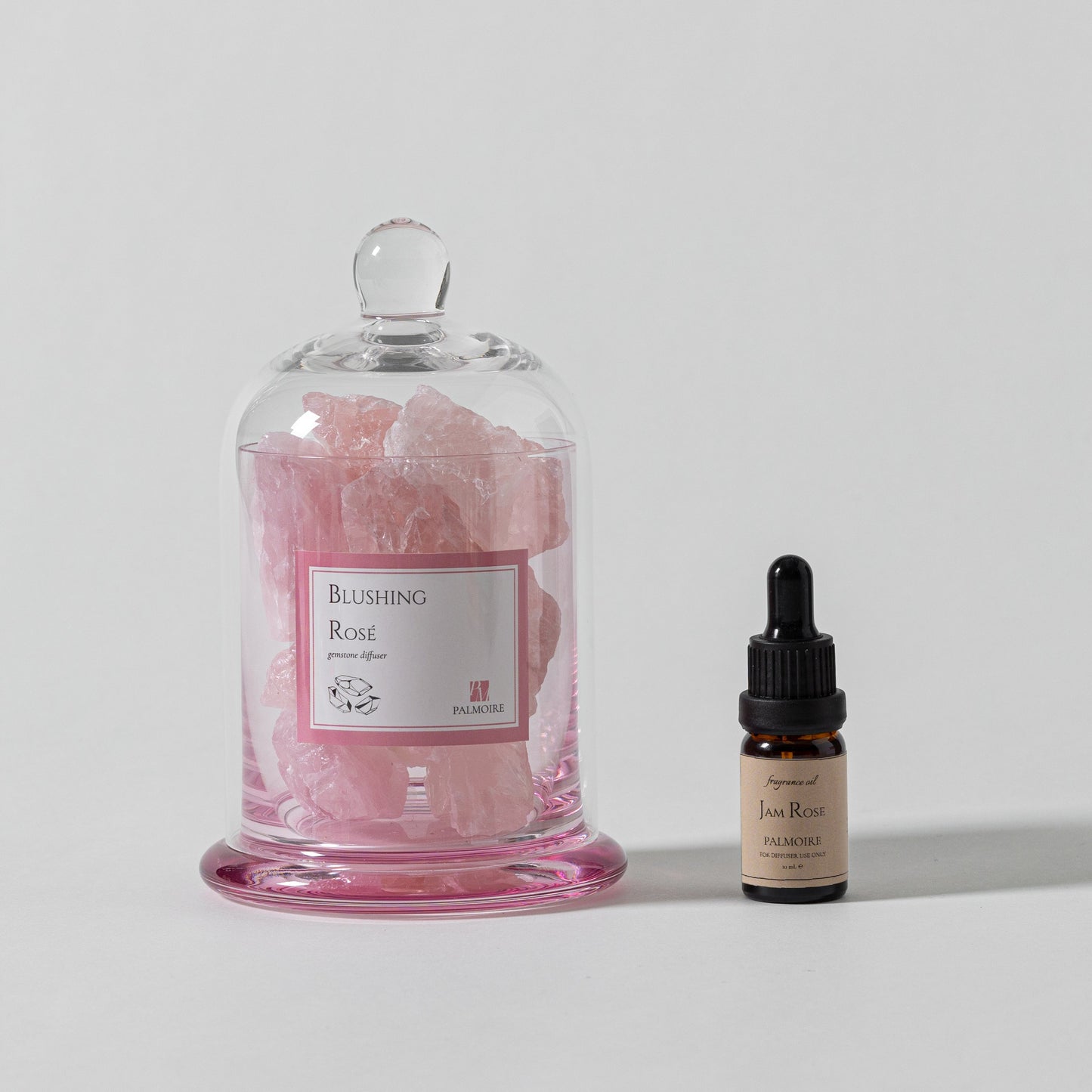 Pink floral product in a glass dome with a small bottle on a light gray background