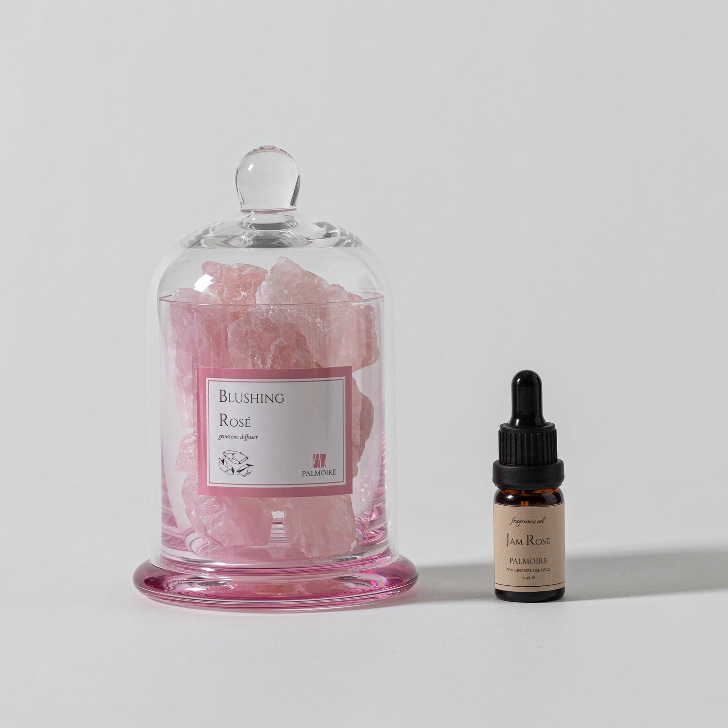 Pink floral product in a glass dome with a small bottle on a light gray background