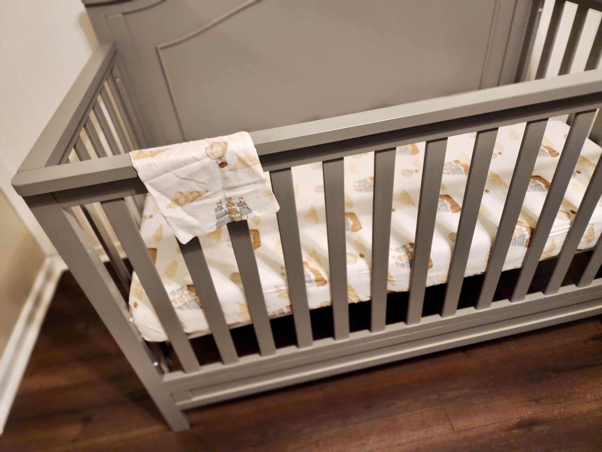Gray crib with a patterned white mattress cover on a wooden floor.