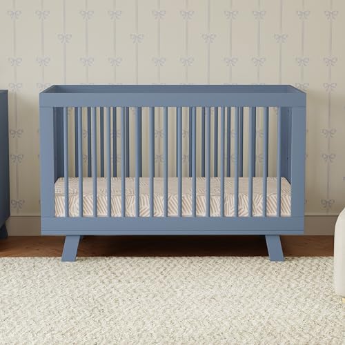Blue crib with a white and gray striped mattress against a wall with decorative wallpaper.