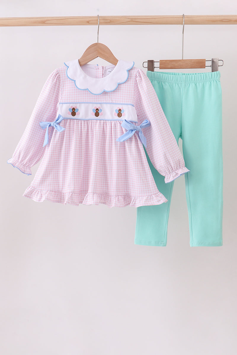 Children's outfit with pink checkered dress and green pants on a wooden hanger against a white background