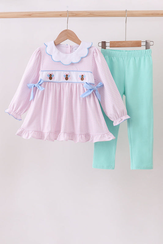Children's outfit with pink checkered dress and green pants on a wooden hanger against a white background