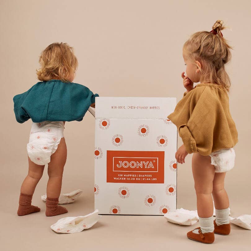Two children standing next to a box of Joonya diapers on a beige background.