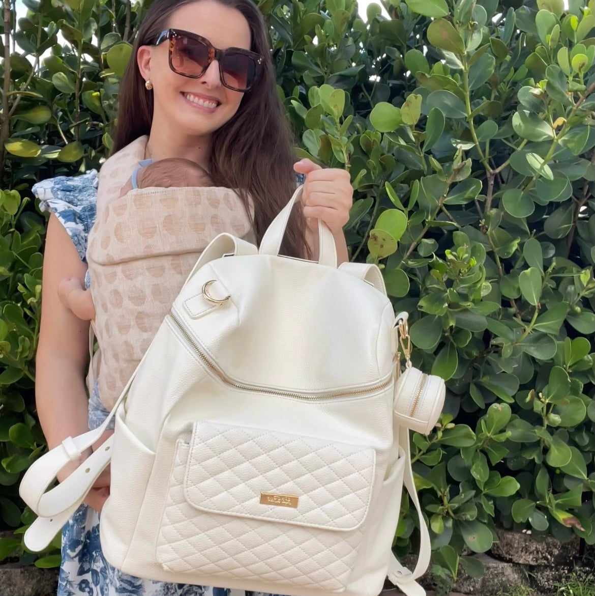 Woman holding a white quilted backpack with a baby in a carrier against a green bush background