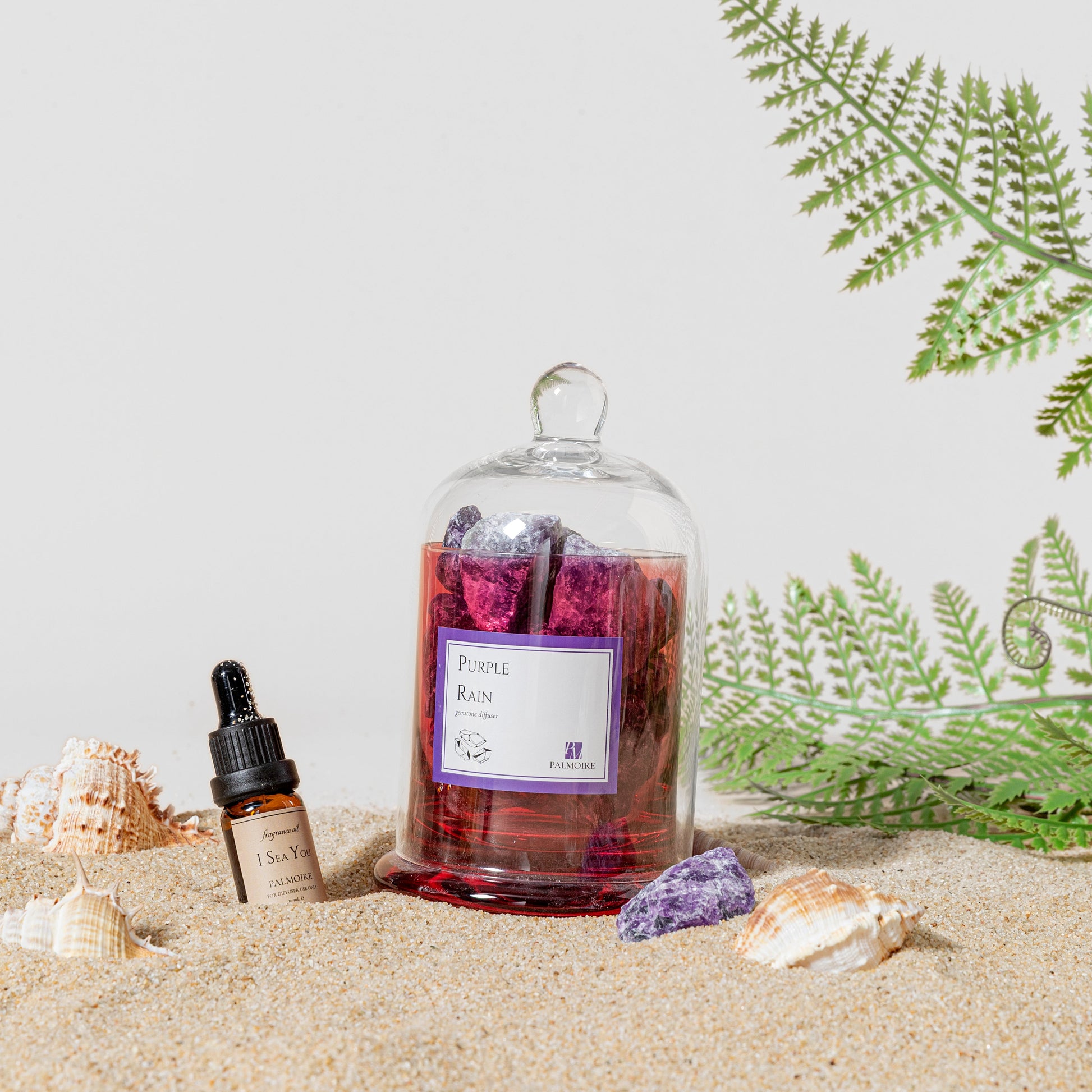 Glass bottle with a purple label on a sandy surface with plants and shells.