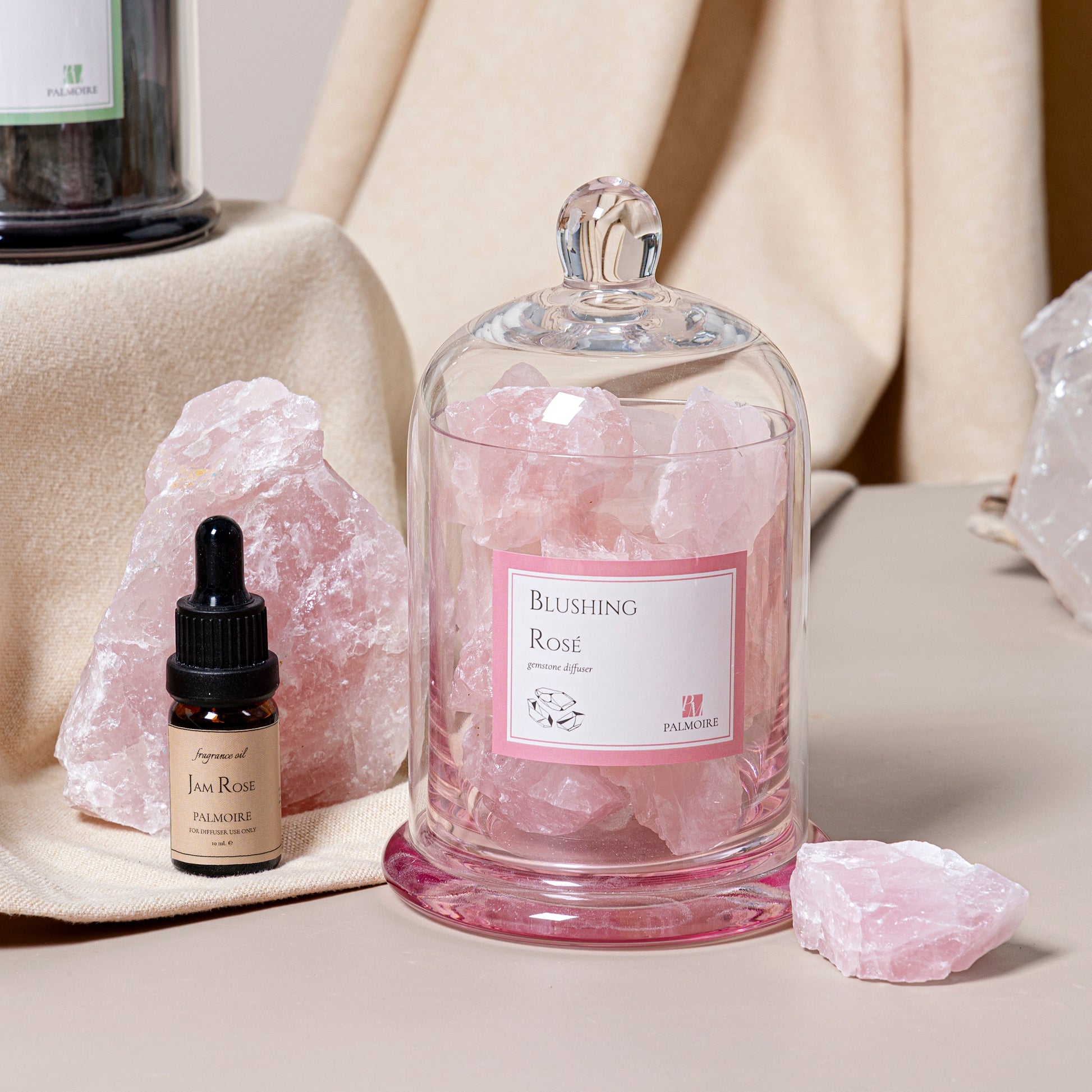 Glass container with pink stones labeled 'Blushing Rose' next to a small bottle on a neutral background