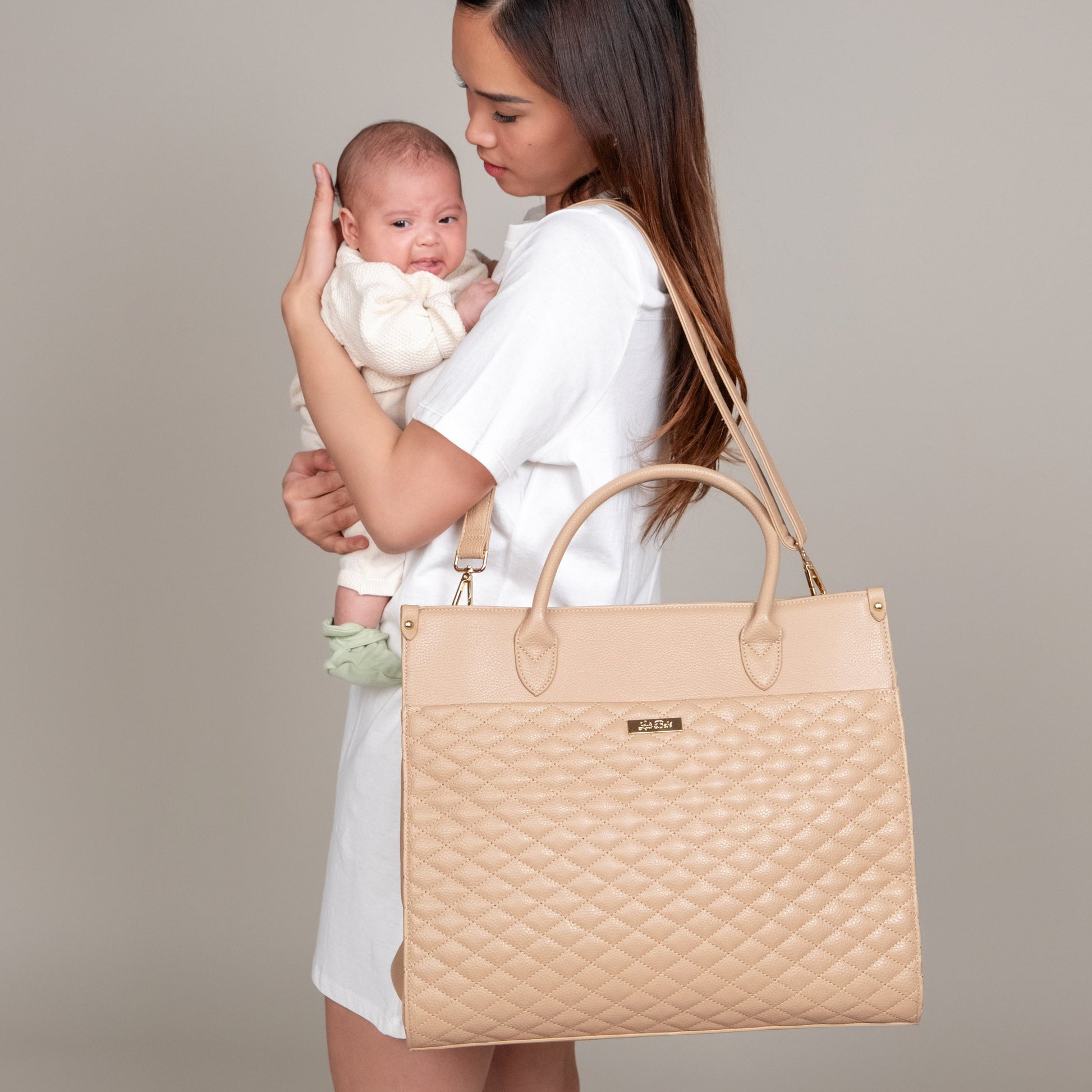 Woman holding a baby and a beige quilted handbag against a neutral background