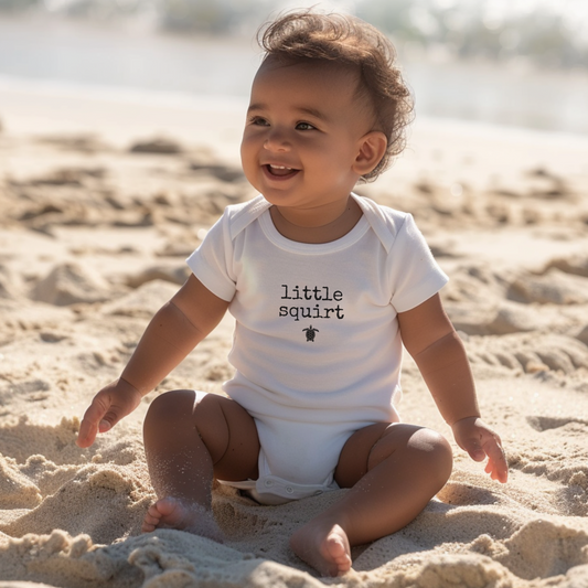 Baby sitting on a sandy beach wearing a 'little squirt' onesie.
