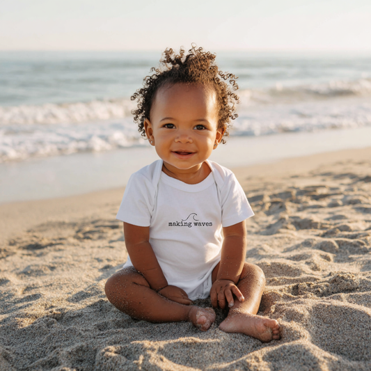 Baby sitting on the beach wearing a white onesie with text.