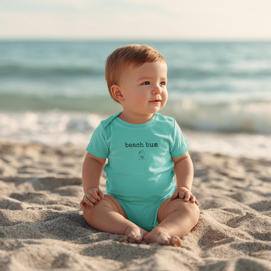 Baby in a turquoise 'beach bum' onesie sitting on a sandy beach with ocean in the background