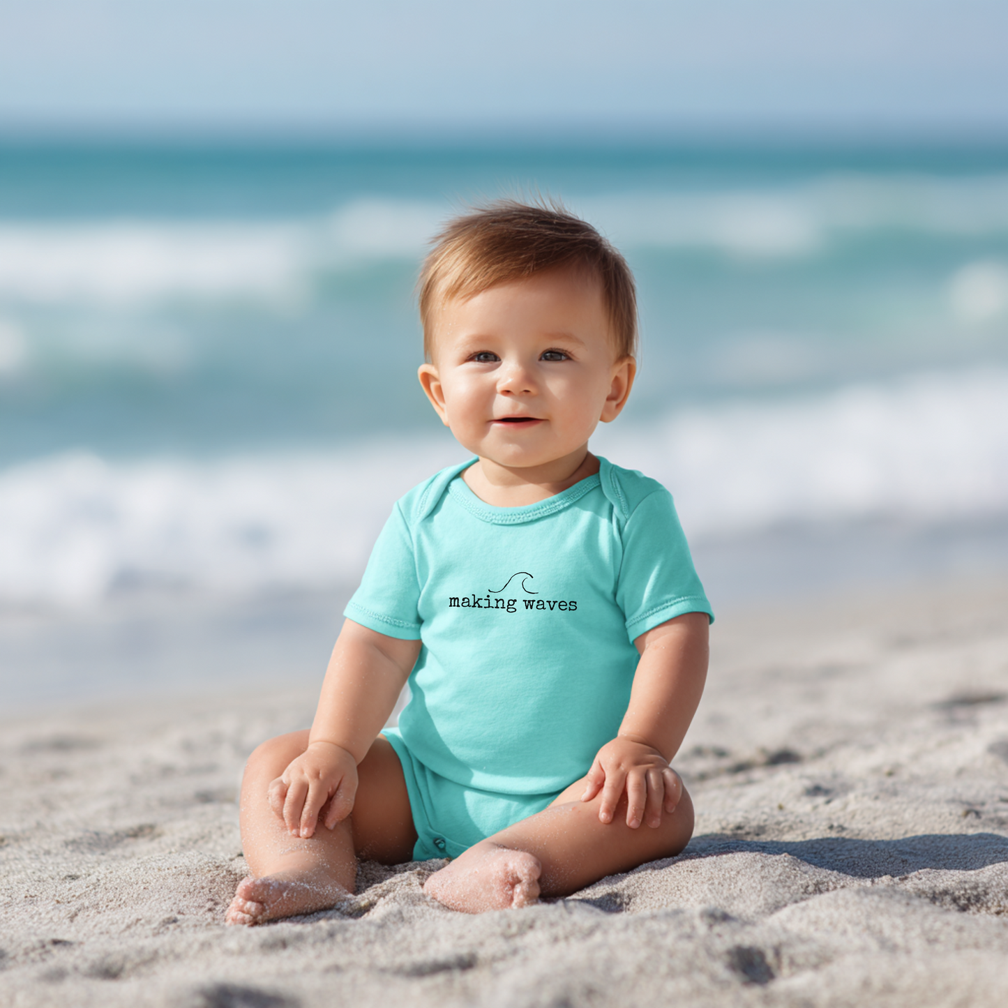 Baby sitting on a beach wearing a turquoise onesie with 'making waves' text.