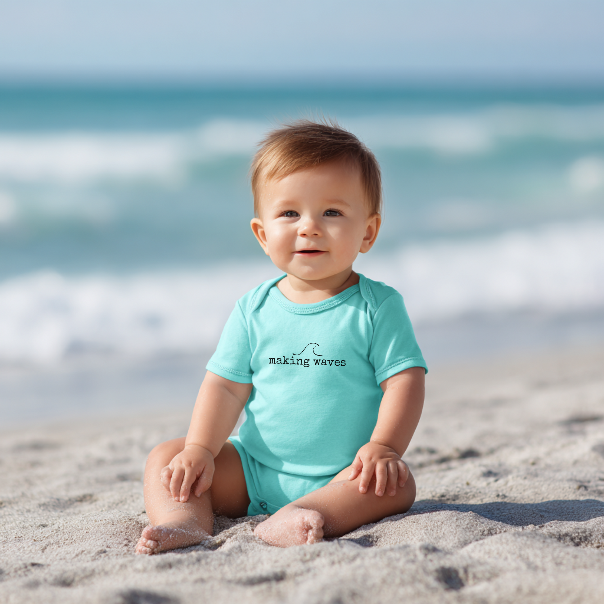 Baby sitting on a beach wearing a turquoise onesie with 'making waves' text.