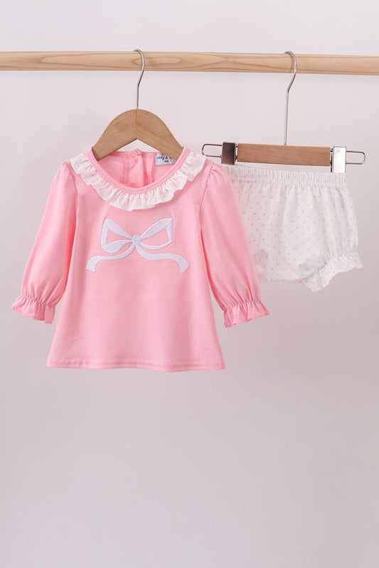 Pink children's top with bow design and white shorts on a wooden hanger against a light gray background