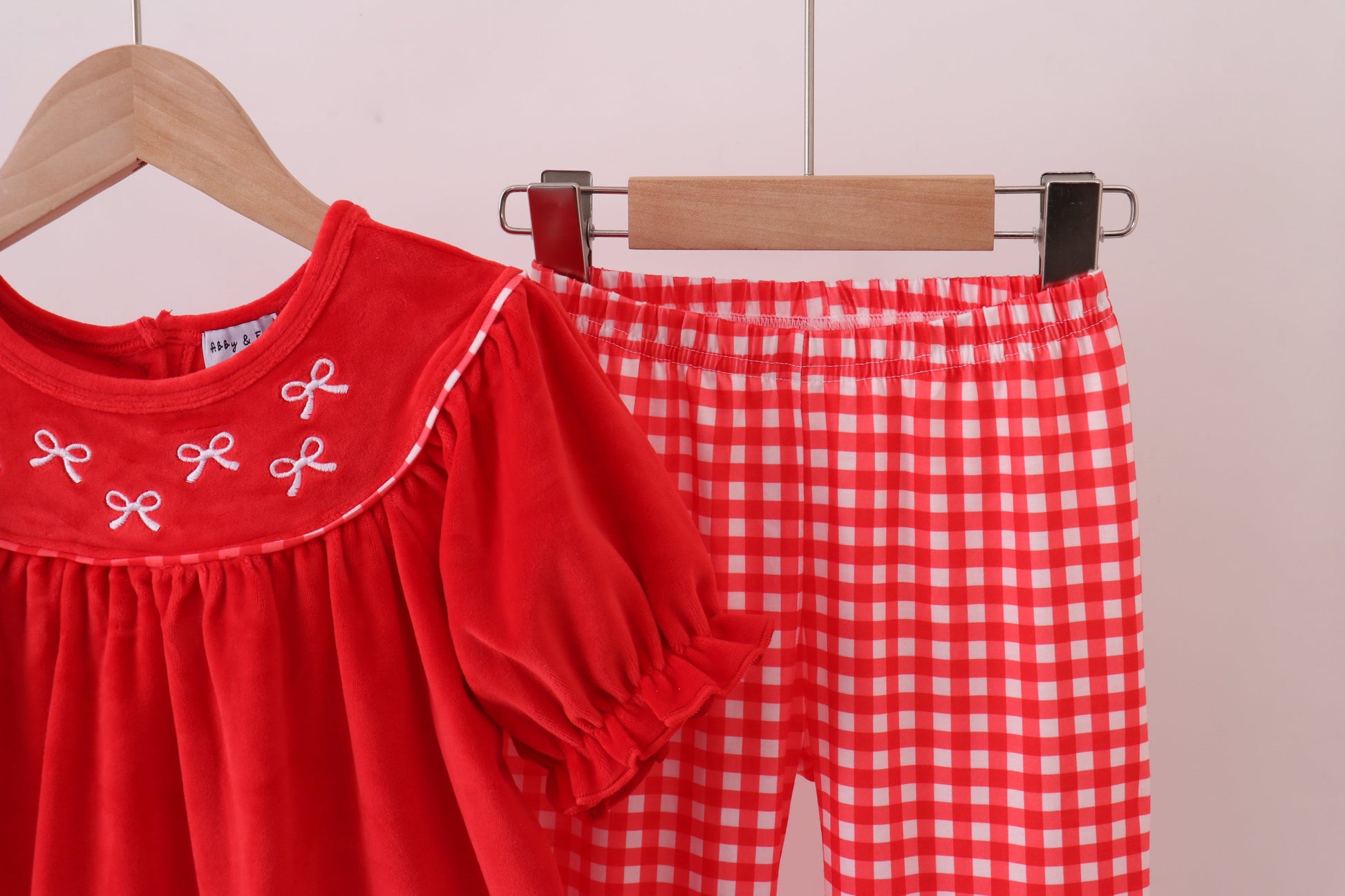 Red children's outfit with white embroidery and red checkered pants on a wooden hanger against a light background.