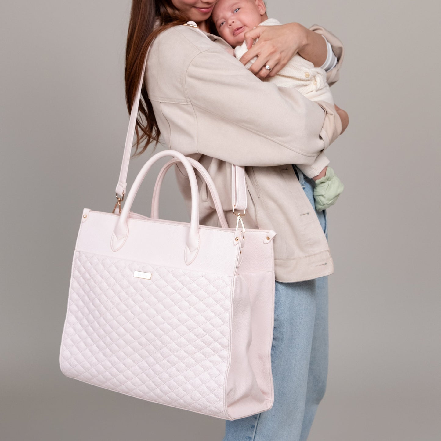 Woman holding a baby and a white quilted handbag against a gray background