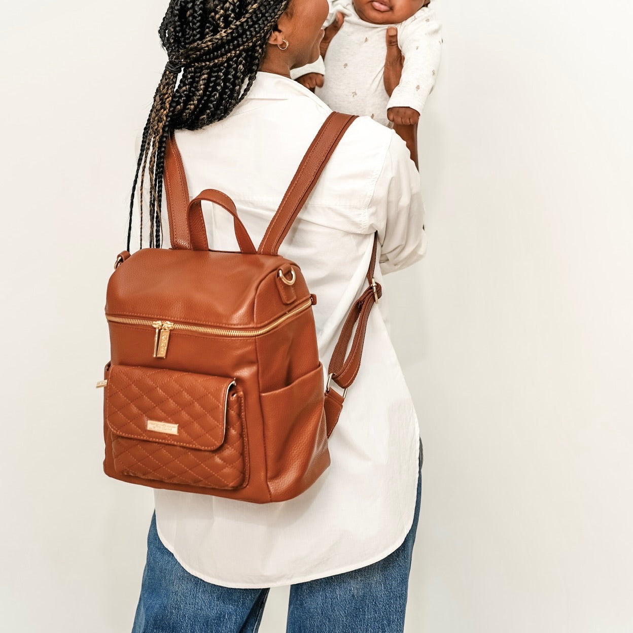 Woman carrying a brown backpack with a baby in a white outfit on her arm against a plain background