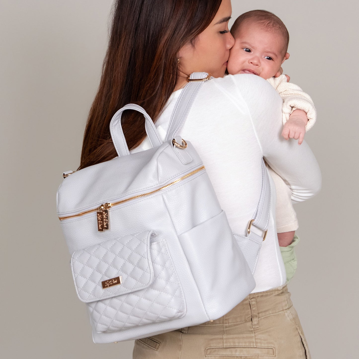 Woman carrying a baby and a white quilted diaper bag against a neutral background