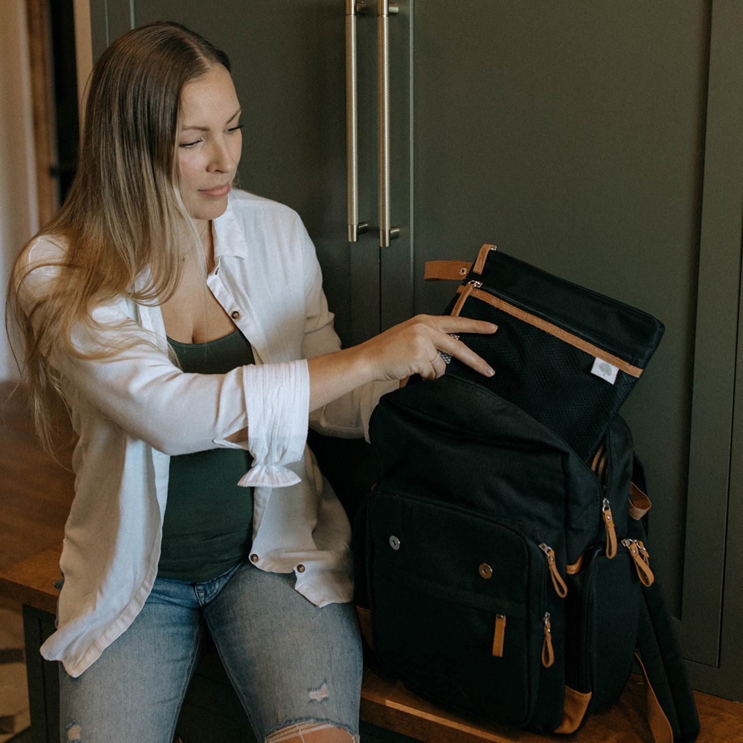 Woman pointing at a black backpack with brown zippers in a kitchen setting.