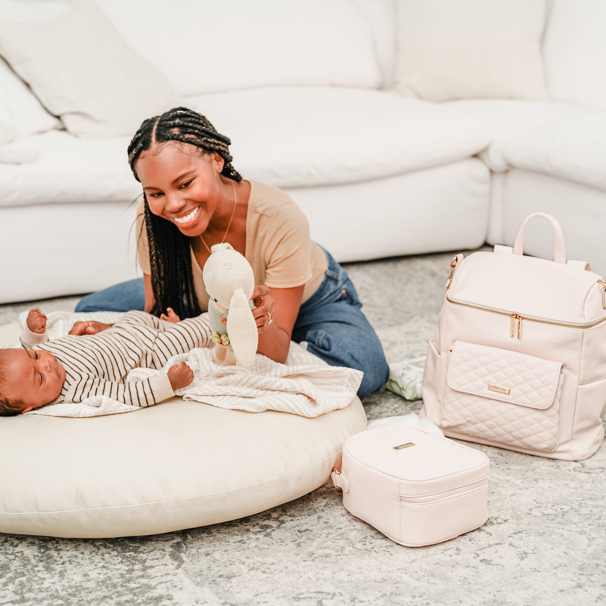 Woman playing with a baby on a white floor with a pink backpack and bag in the foreground.