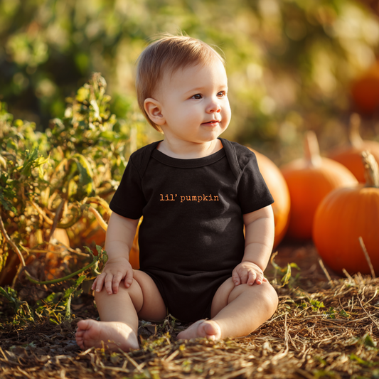Baby wearing a black onesie with 'lil' pumpkin' text, sitting in a pumpkin patch.