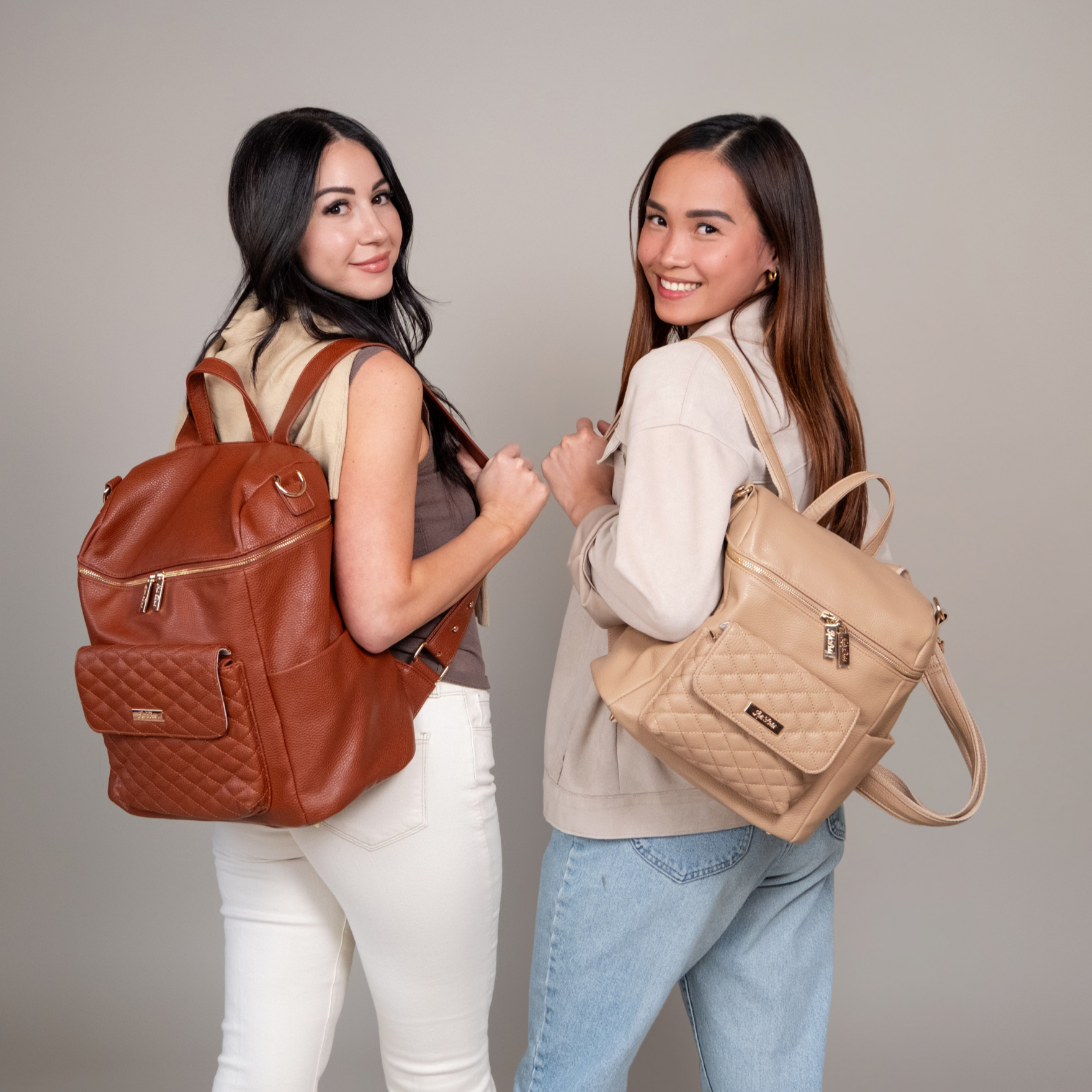 Two women holding a brown backpack and a beige backpack against a plain background