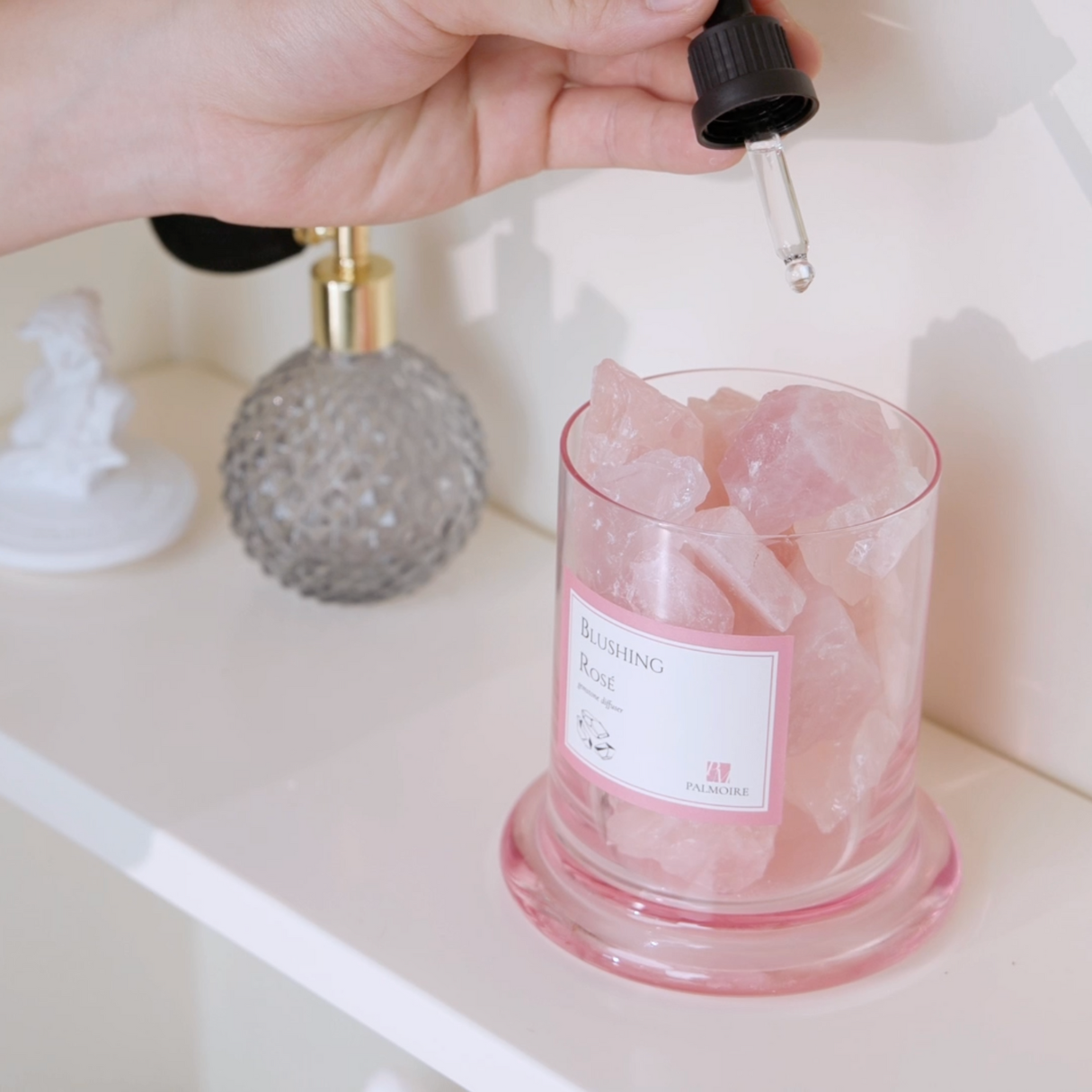 Hand dropping a liquid into a pink glass container labeled 'Bath Salt' with a dropper.